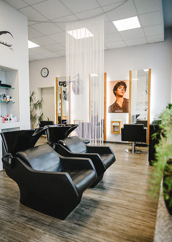 Moderner, heller Innenbereich des Salon Haarscharf mit zwei schwarzen Haarwaschsesseln im Vordergrund und Bedienplätzen mit Spiegeln im Hintergrund.
